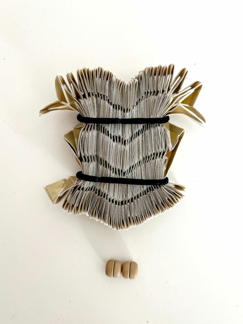 Un libro abierto e intrincadamente doblado de la colección 3Dtachables se sujeta mediante correas negras sobre dos pares de pequeños objetos redondos de color beige sobre una superficie blanca.