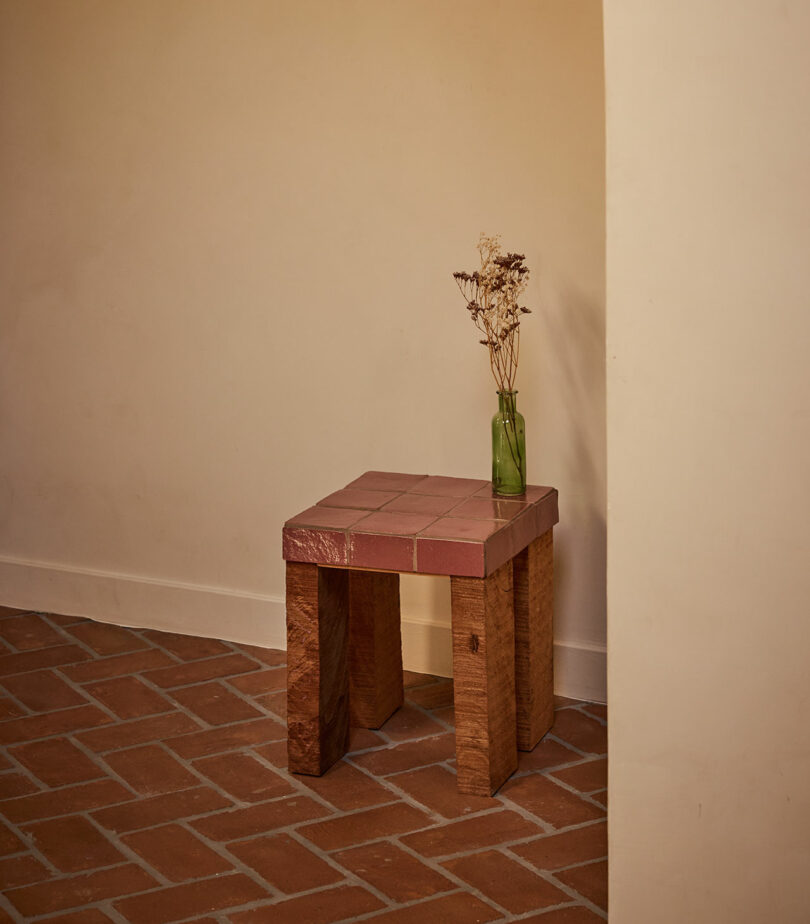 A small wooden table with a pink tiled top from Ah Um holds a green glass bottle with dried flowers, placed on a terracotta tiled floor against a beige wall.