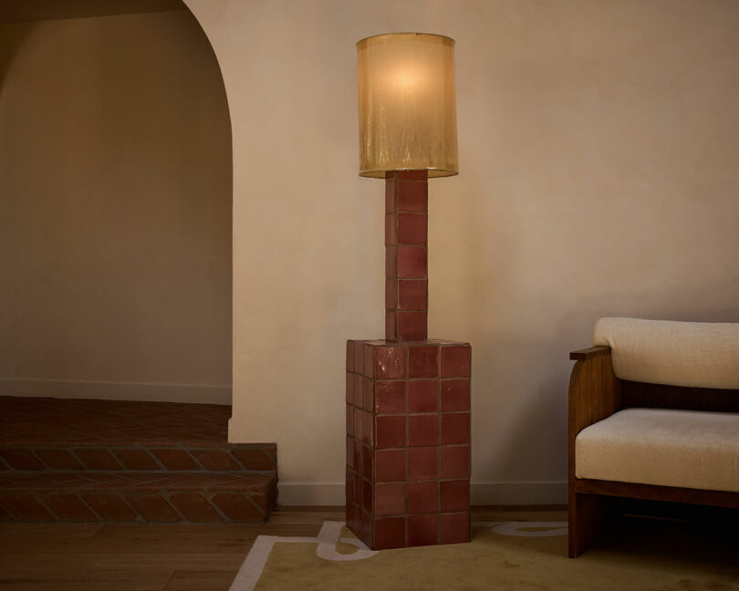 A floor lamp with a square, red-tiled base and a cylindrical beige lampshade stands next to a wooden armchair in a softly lit room with Ah Um charm, featuring an arched doorway and tiled steps.