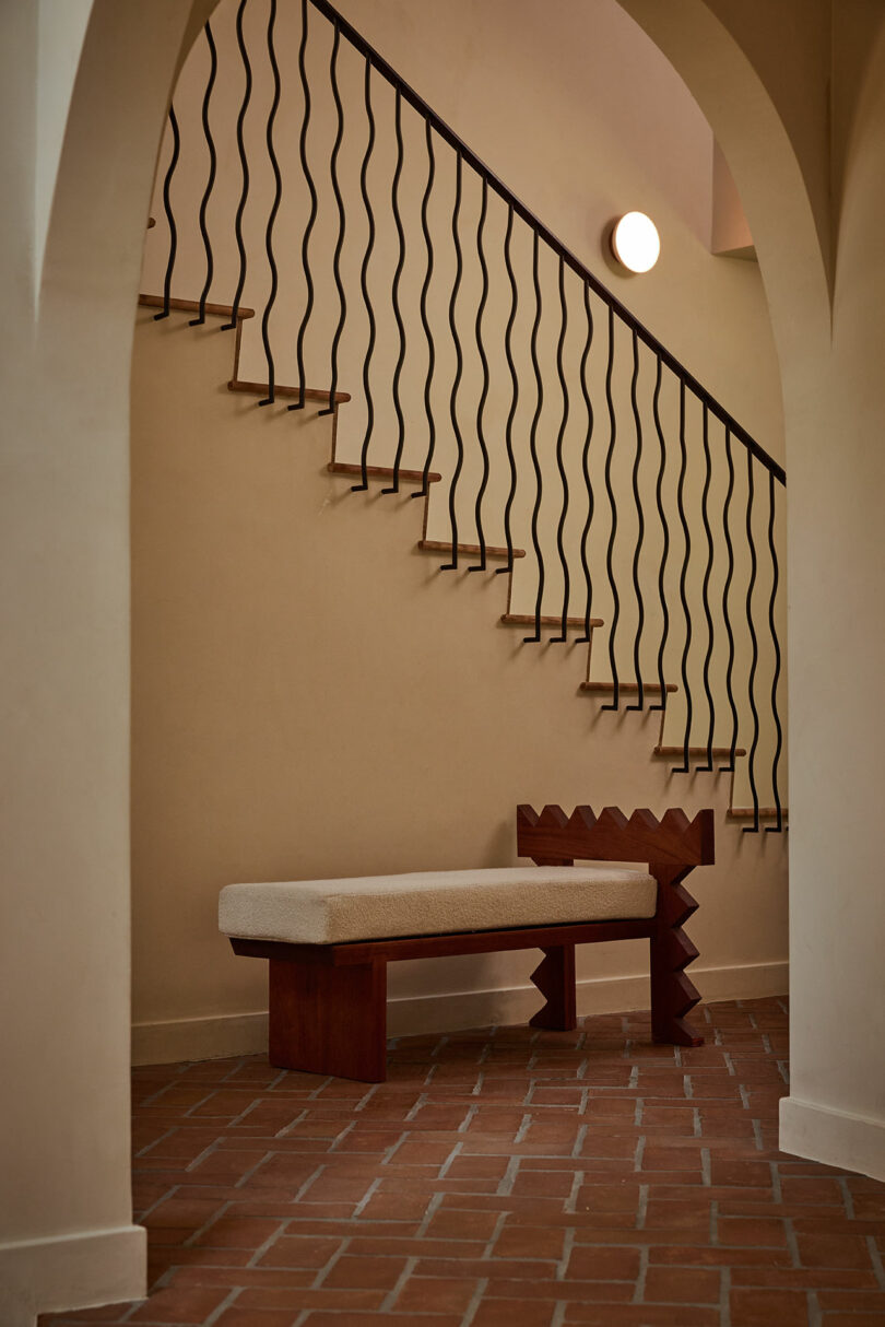 A modern hallway with a zigzag-back wooden bench by Ah Um, terracotta tile flooring, arched entryway, and a staircase with wavy black metal railing and a round wall light.