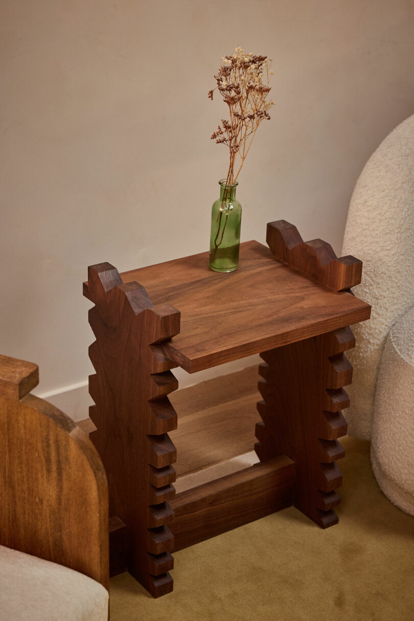 A small wooden side table with decorative stepped edges from Ah Um holds a green glass vase with dried flowers, placed between two upholstered chairs.