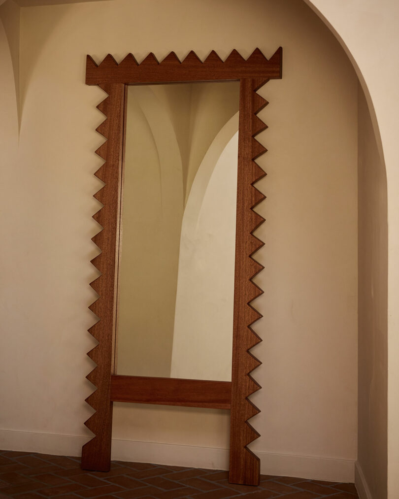 A tall wooden-framed mirror from Ah Um with a zigzag edge stands against a cream-colored wall and arched doorway on a tiled floor.