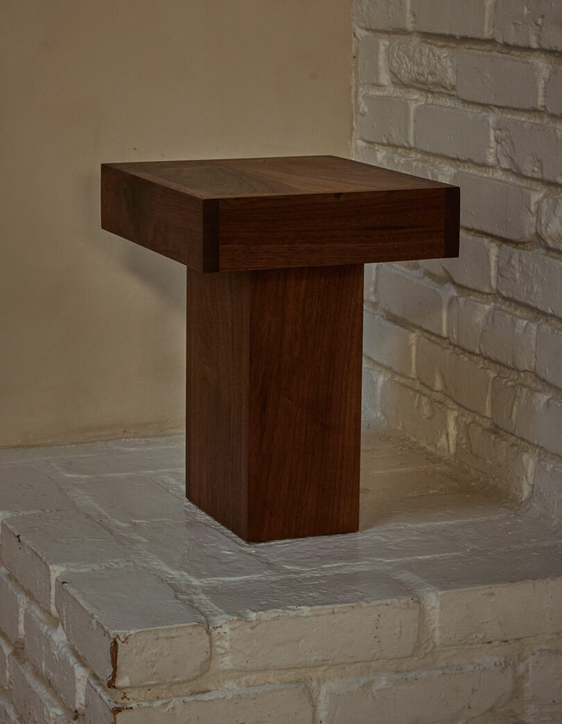 A rectangular wooden pedestal with a wide top and narrow base, reminiscent of the Ah Um style, is placed on a white-painted brick hearth next to a white brick wall.