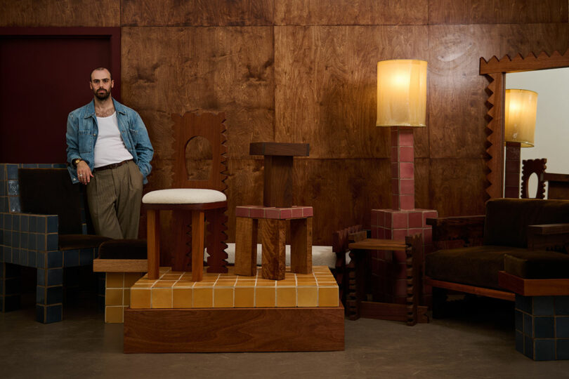 A man in a denim jacket stands in a wood-paneled room with Ah Um geometric, sculptural furniture and a large mirror. Lamps and chairs feature tile patterns in earthy tones.