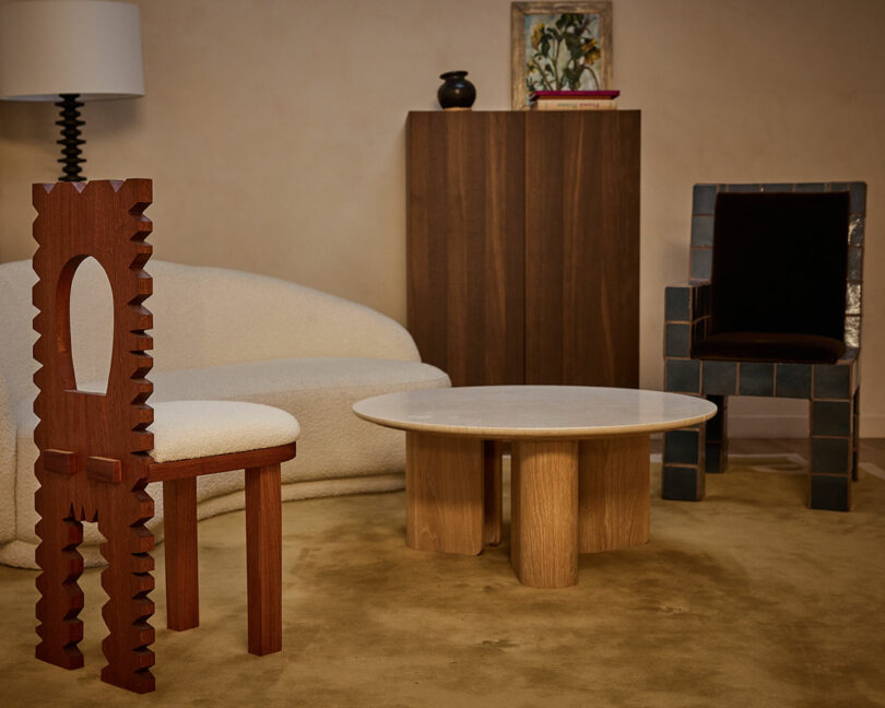 A living room with a sculptural Ah Um wooden chair, a curved white sofa, a round wooden coffee table, a dark armchair, a wooden cabinet, a lamp, and artwork on the wall.