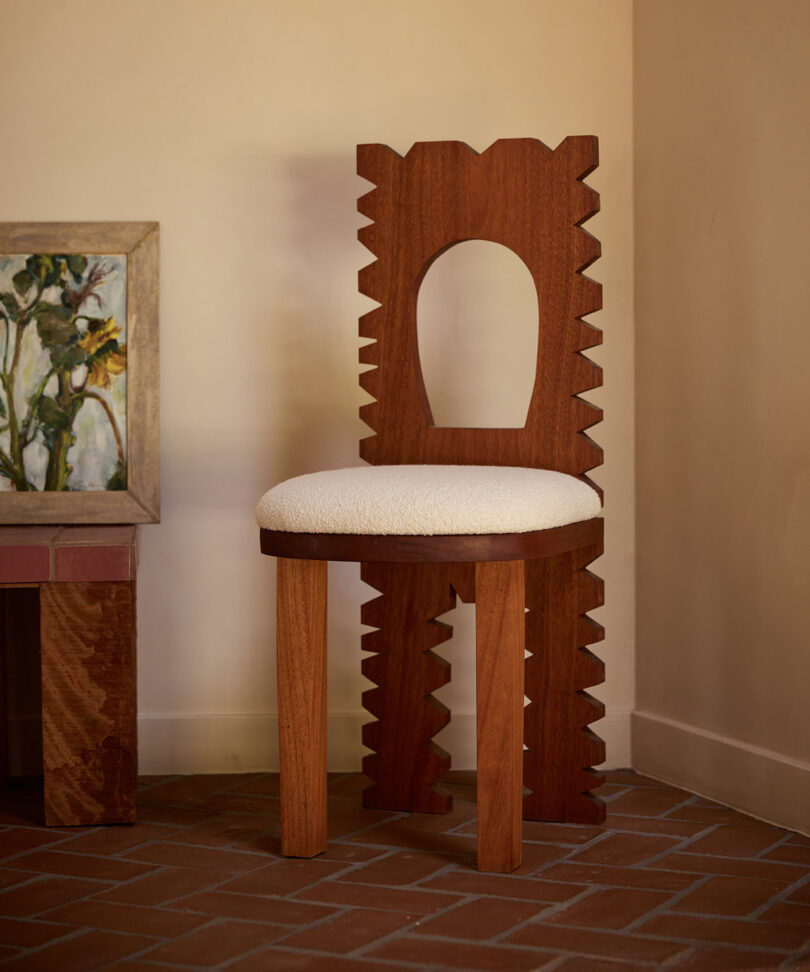 A wooden chair from Ah Um with a sculpted back featuring zigzag edges and a round cutout, upholstered seat, sits on a tiled floor next to a framed painting.
