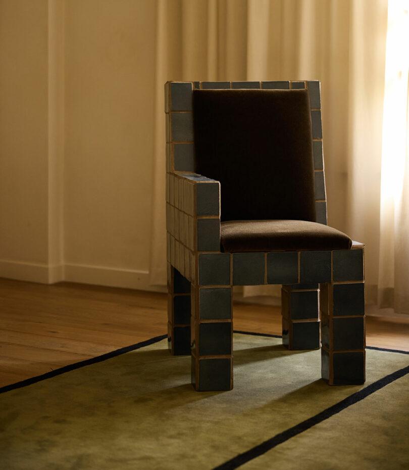 A chair with a dark cushion and square, tiled Ah Um frame sits on a green rug in a softly lit room with wooden flooring and light curtains.
