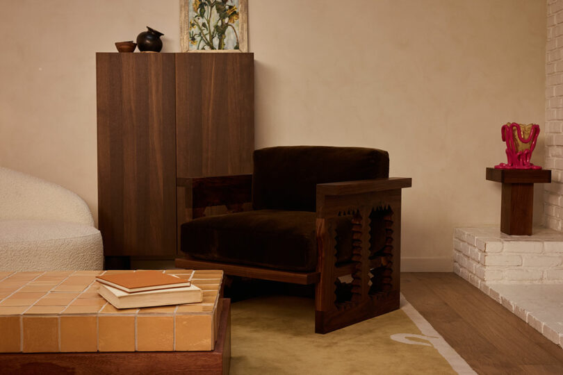 A living room with a dark wooden armchair, cabinet, tiled coffee table with books, small side table with a pink Ah Um sculpture, and artwork against a beige wall.