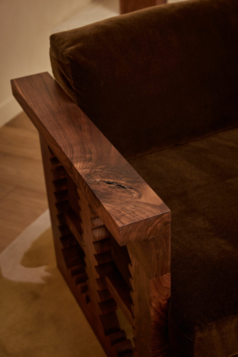 A close-up of the Ah Um brown velvet armchair with a wooden armrest, featuring a natural knot and geometric cutout design.