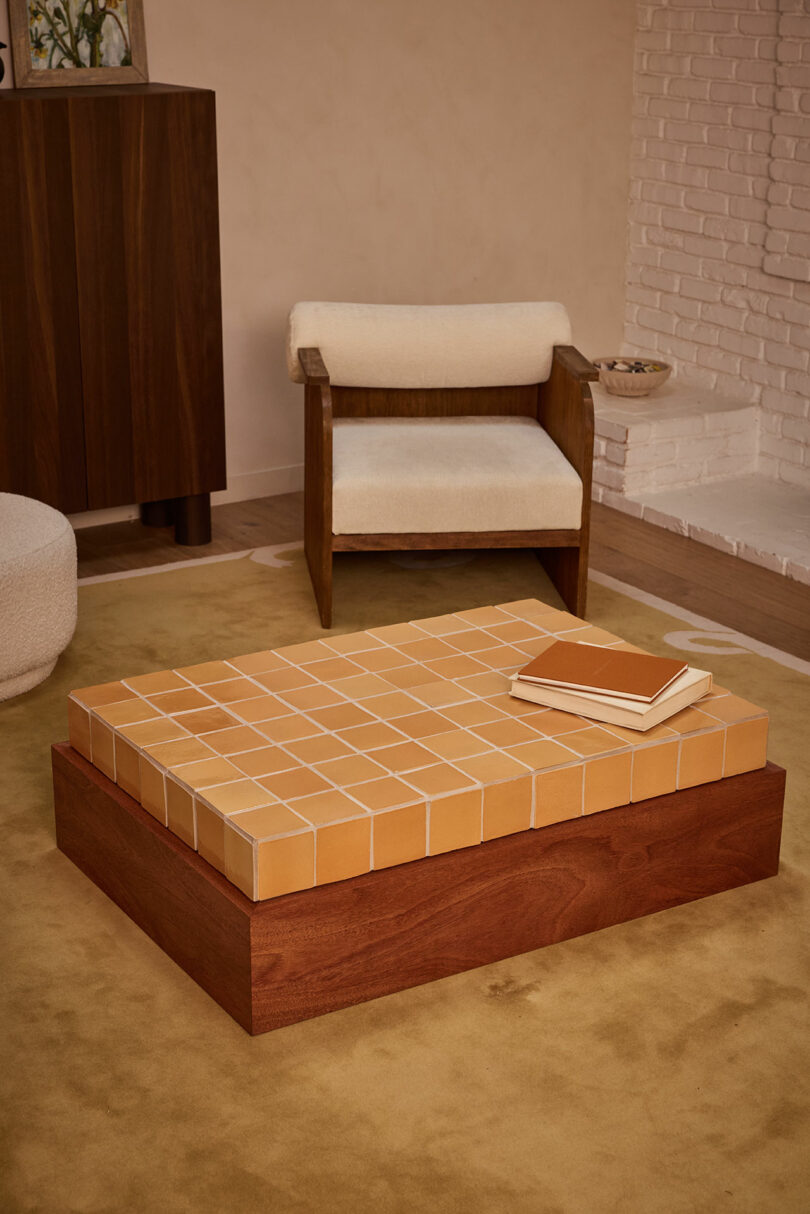 A wooden coffee table with a tiled surface, two closed books on top—one titled "Ah Um"—an armchair with cream cushions, and a yellow rug in a cozy living room setting.