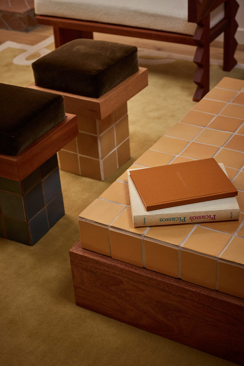A tiled coffee table with two books and a notebook on top is surrounded by cushioned stools, a yellow rug, and the relaxed ambiance of Ah Um.