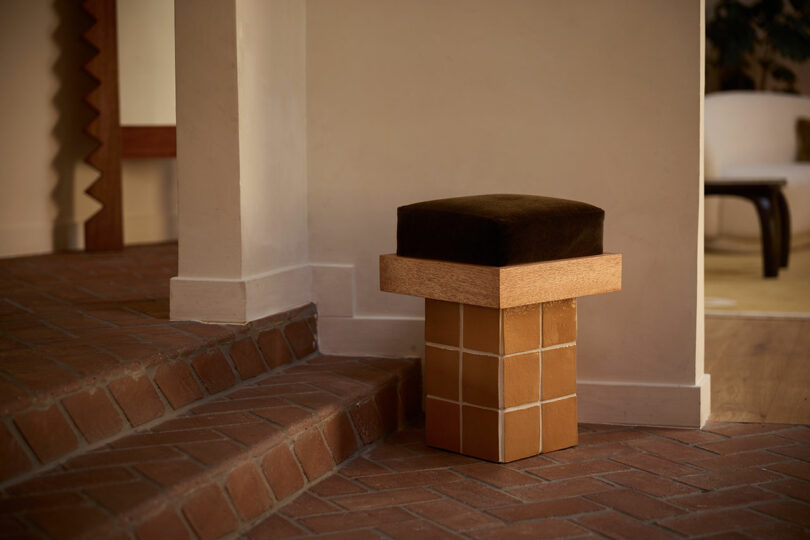 A geometric stool with a square, brown cushioned seat and a tiled cube base from Ah Um sits next to brick steps and a beige wall in a modern interior.