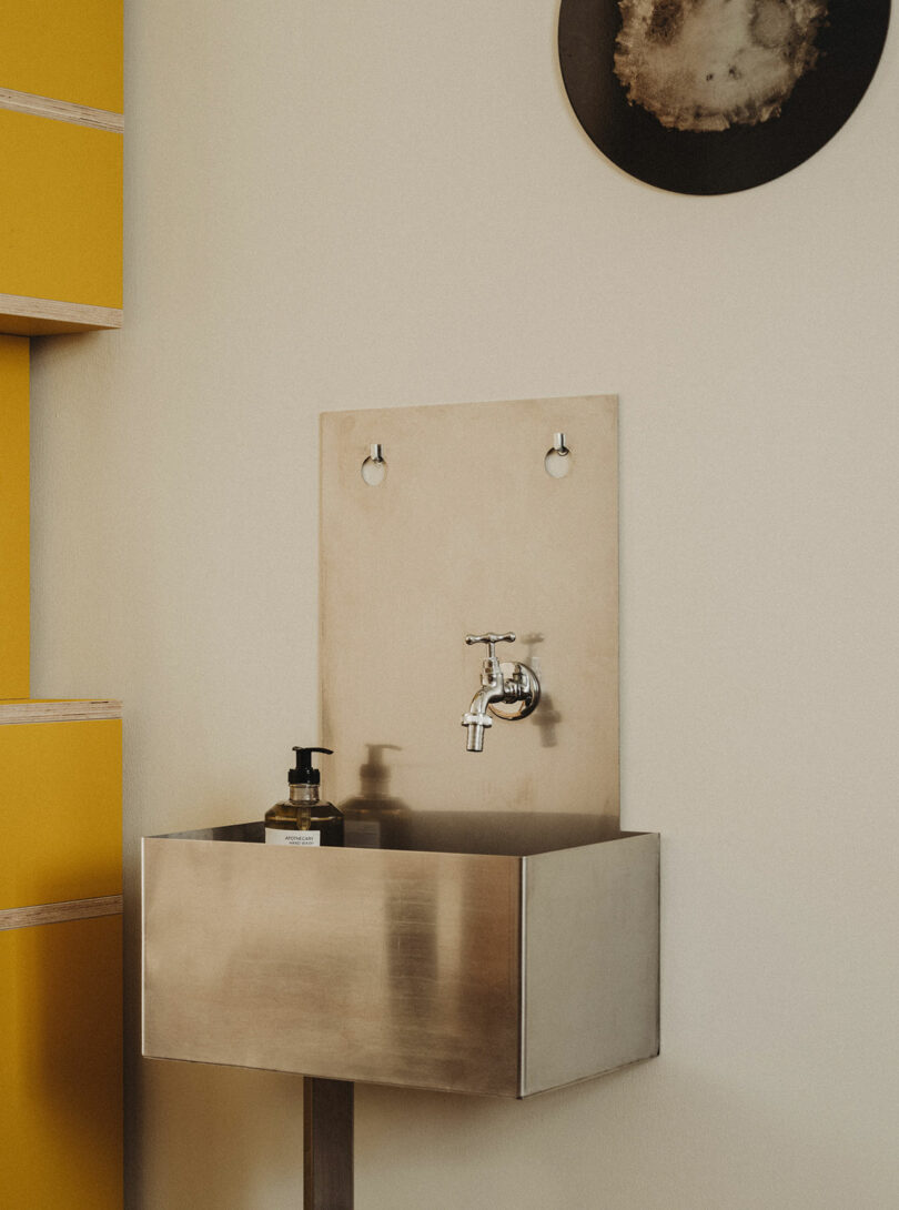 A stainless steel sink with a wall-mounted faucet, soap dispenser, and a beige backsplash, beside yellow shelves and a round wall decoration.
