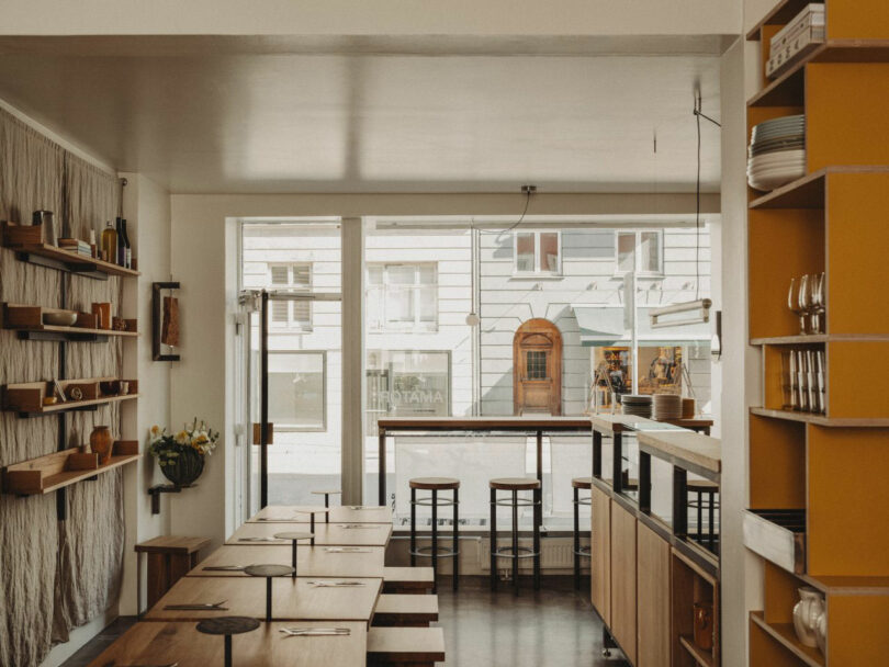 Modern cafe interior with wooden tables, shelves, and stools by large windows facing the street. Natural light fills the space, highlighting minimalistic and cozy decor.