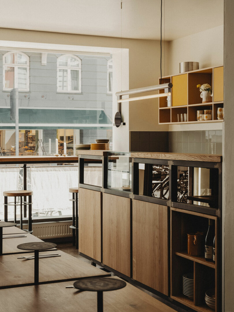 Modern café interior with wooden furniture, open shelving with yellow accents, and large windows overlooking a street with buildings.
