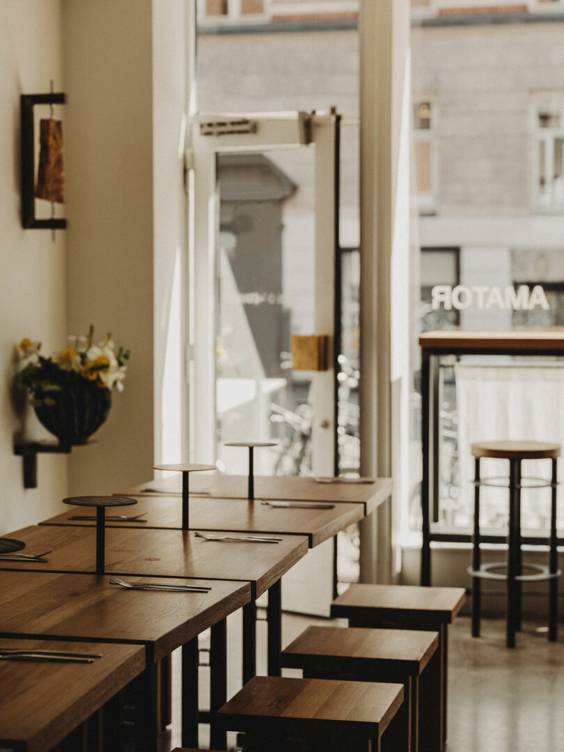 Sunlit café interior with wooden tables and stools, neatly set with cutlery; large windows offer a view of the street and buildings outside.