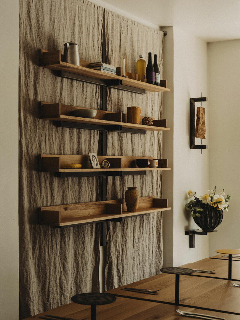 Wooden shelves holding books, bottles, vases, and décor items are mounted on a wall draped with beige fabric above a wooden dining table set for three.