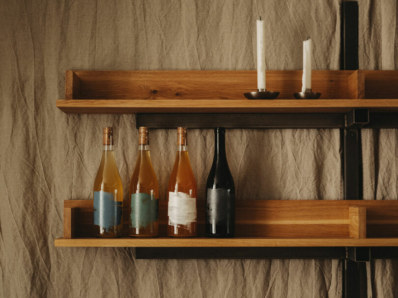 Four wine bottles with minimalist labels are displayed on a wooden shelf, next to two partially burned white candles in holders against a beige fabric background.