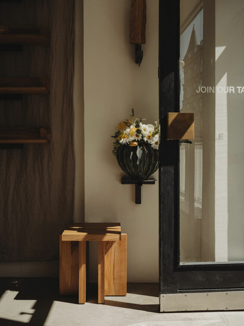 A wooden stool sits beside a glass door with a gold handle; a vase of white and yellow flowers is mounted on the wall above the stool.