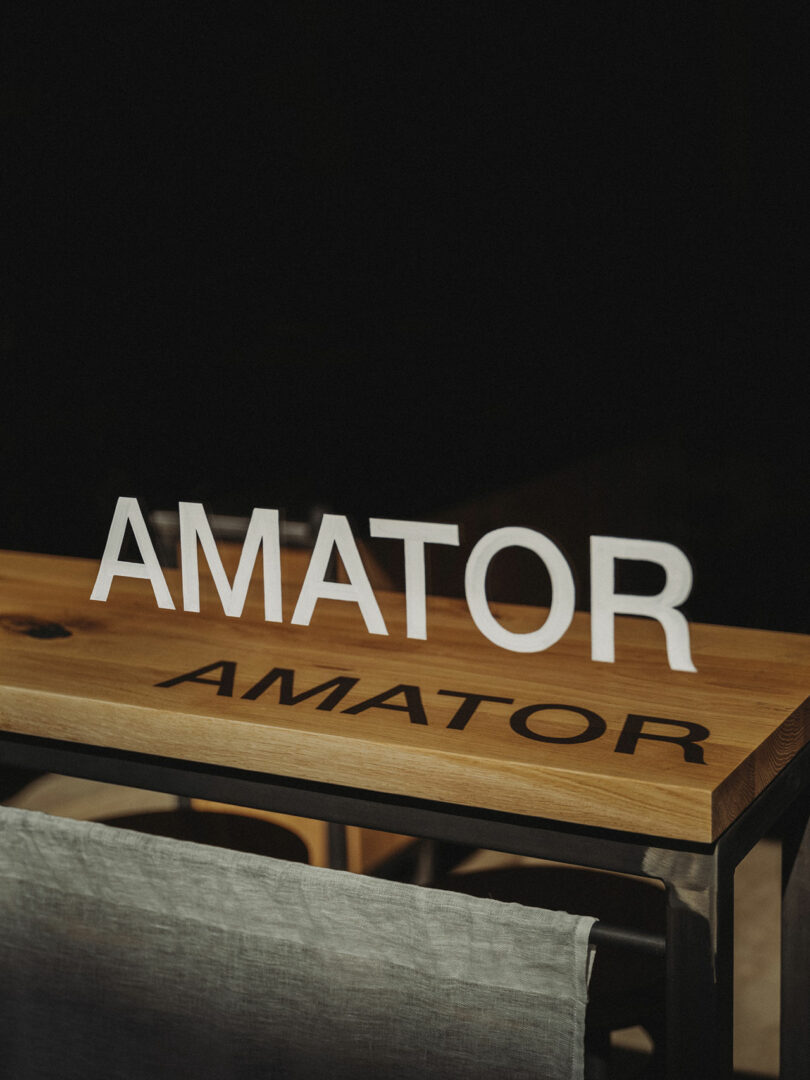 A wooden table with the word "AMATOR" in 3D white letters standing on top and the same word printed in black on the table surface.