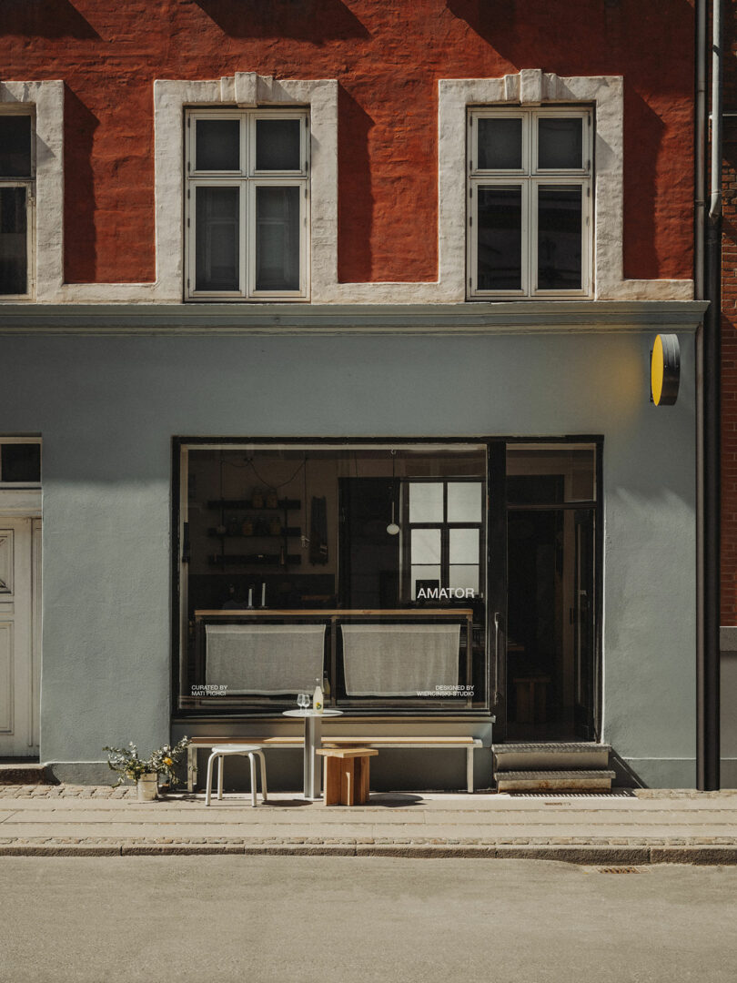 A small café with a blue and red exterior, large windows, and a bench with stools and a table outside on the sidewalk.