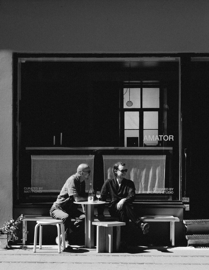 Two people sit at a small outdoor table in front of a café window, engaged in conversation on a sunny day.
