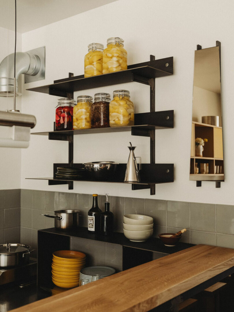 Modern kitchen shelves with jars of preserved food, plates, bowls, and utensils arranged neatly above a countertop with cookware and condiments.