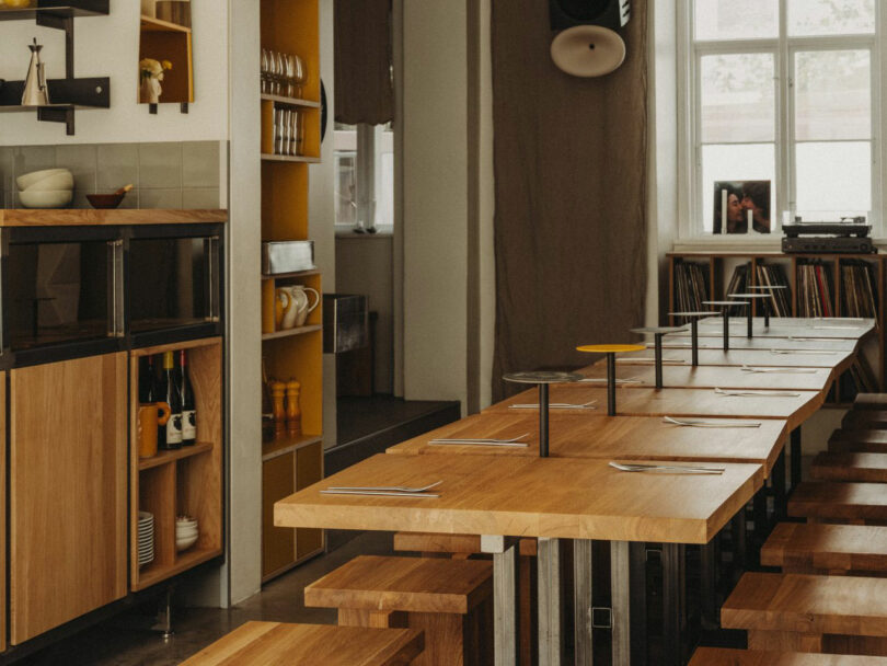 A modern dining area with wooden tables and benches is set with utensils; shelves and cabinets hold dishes, bottles, and kitchenware under natural light from a large window.