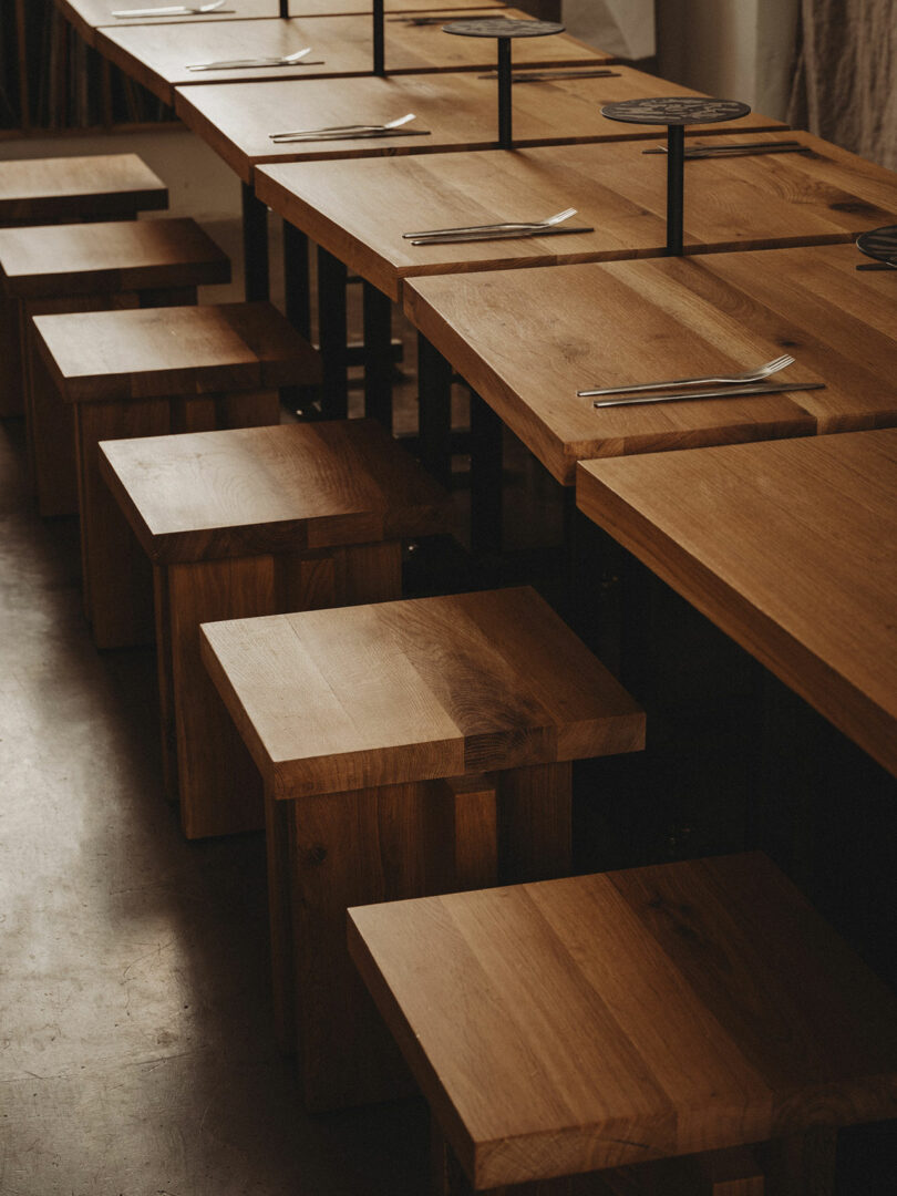 A row of wooden tables and square stools set with forks and knives in a minimalist indoor dining area.