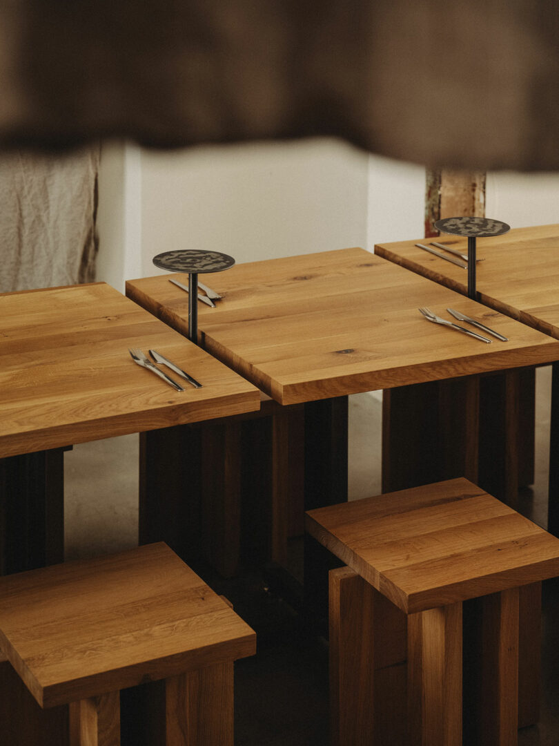 Wooden tables with matching benches, each set with a fork and knife, are arranged in a minimal, modern dining space with neutral-toned walls.