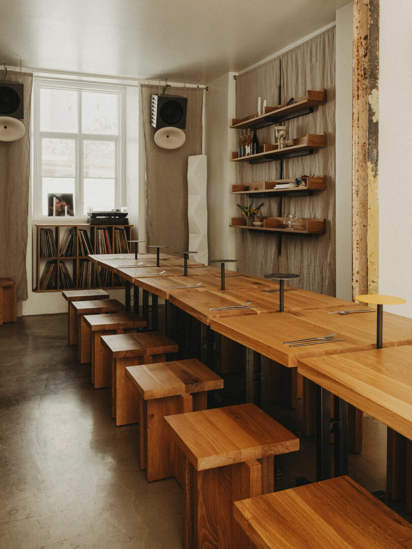 A minimalist dining area with long wooden tables and stools, shelves with decor, speakers, and a window letting in natural light.