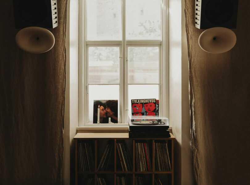 A window above a shelf holding vinyl records, a record player, speakers, and two album covers, one showing two kissing and the other featuring red illustrated faces.