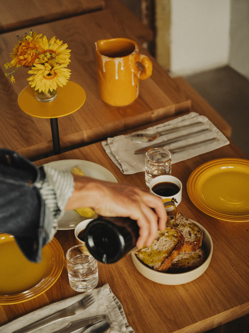 A person pours liquid onto slices of bread on a wooden table set with yellow dishes, glasses of water, utensils, and a small vase of yellow flowers.