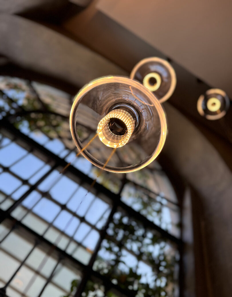Close-up view of a lit Aura Snøhetta modern pendant light fixture, with blurred large windows and lush greenery in the background.