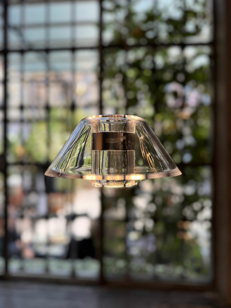 A modern Aura Snøhetta glass pendant light hangs indoors with a large window and blurred greenery visible in the background.
