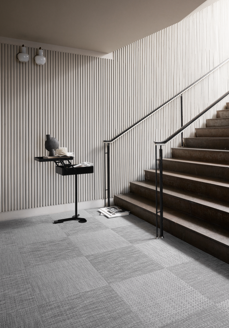 Serene stair landing featuring textured grey carpet tiles, ribbed wall paneling, and a modern black side table with stacked decor.