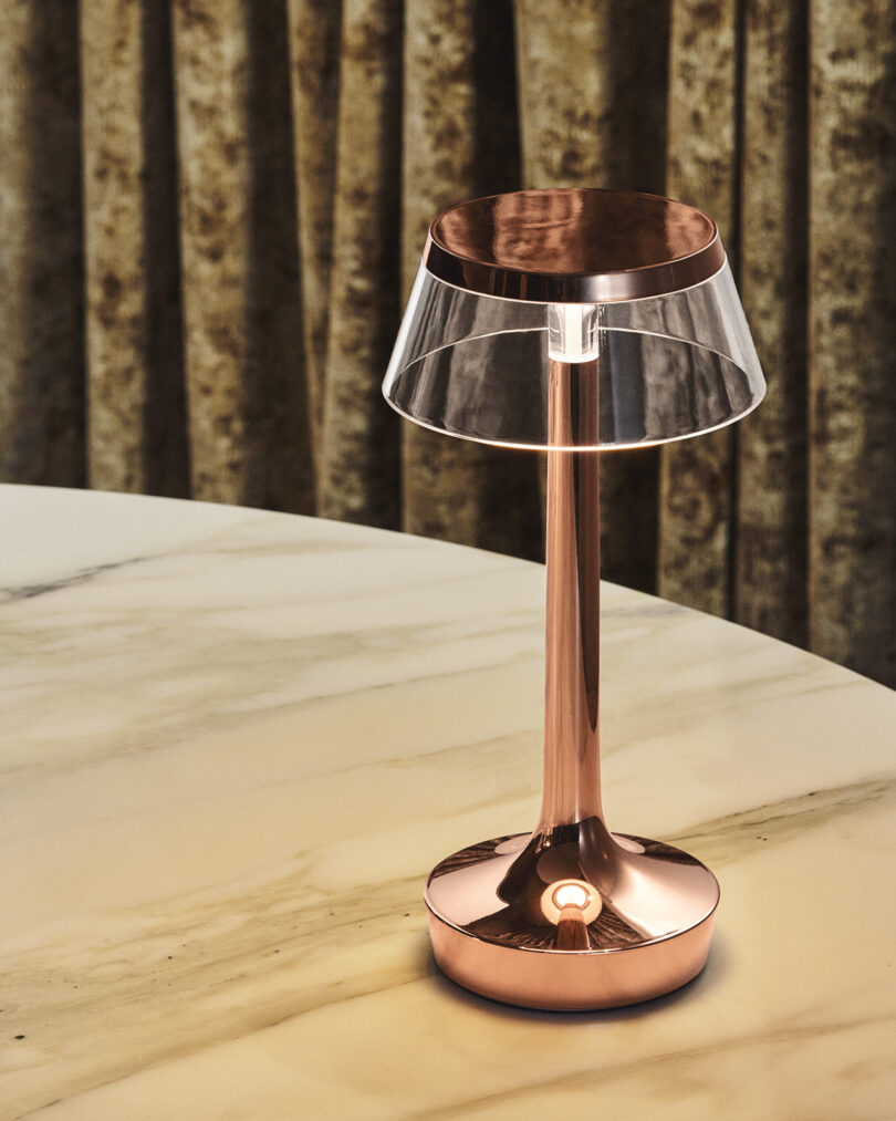 A modern copper table lamp with a clear shade sits on a marble surface, with gold curtains in the background.