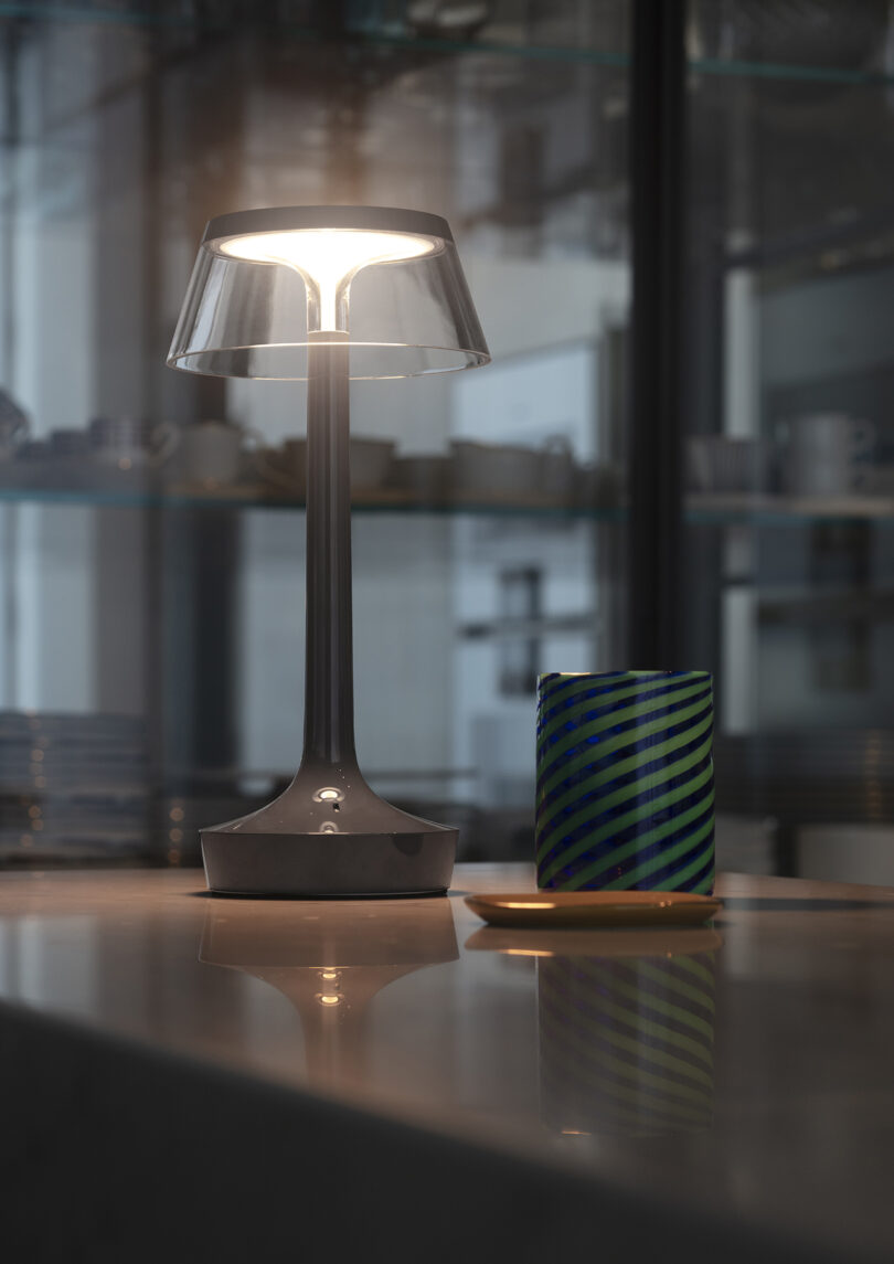 A modern table lamp with a transparent shade is lit on a reflective surface next to a green and blue striped glass and a small plate. Shelves with dishes are visible in the background.