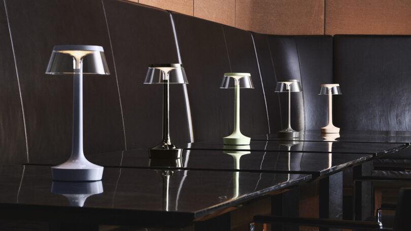 Five modern table lamps in different colors are arranged in a row on a glossy black table with dark leather seating in the background.
