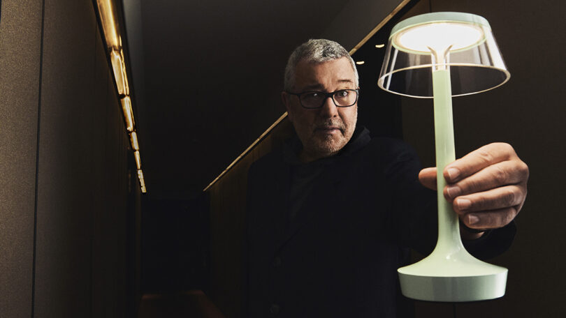 A man with glasses holds a modern table lamp, illuminating his face in a dimly lit hallway.