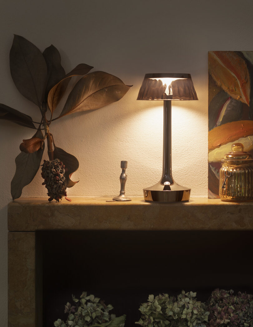 A metallic table lamp is lit on a mantel next to a dried branch, a candlestick, a glass jar, and a painted canvas. Dried flowers are visible in the lower part of the image.