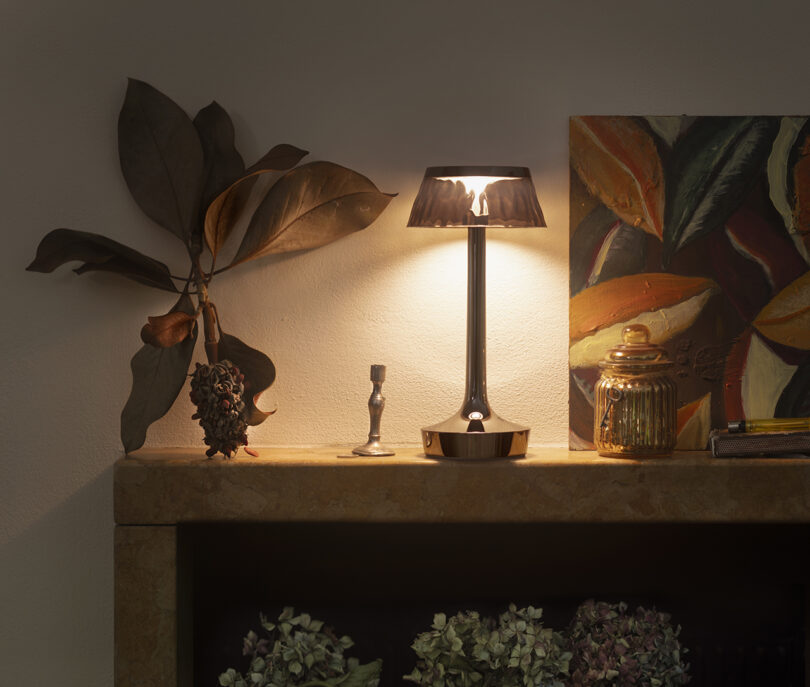 A lit table lamp on a stone mantel, accompanied by dried leaves, a small figurine, a glass jar, and a colorful painting of leaves in the background.