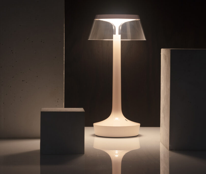 A modern white table lamp with a clear lampshade is illuminated on a glossy surface, surrounded by rectangular and square concrete blocks.