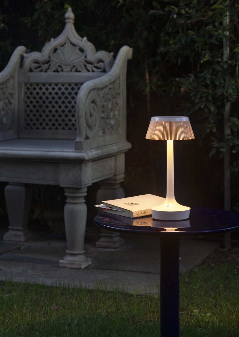 A small white lamp and two books rest on a round dark table outdoors, with an ornate stone bench and greenery in the background.