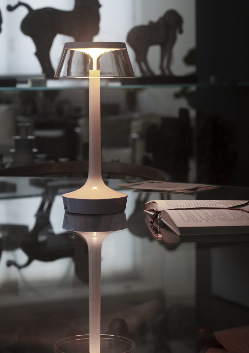 A modern table lamp is illuminated on a glass surface near an open book; animal figurines are visible on a shelf in the background.