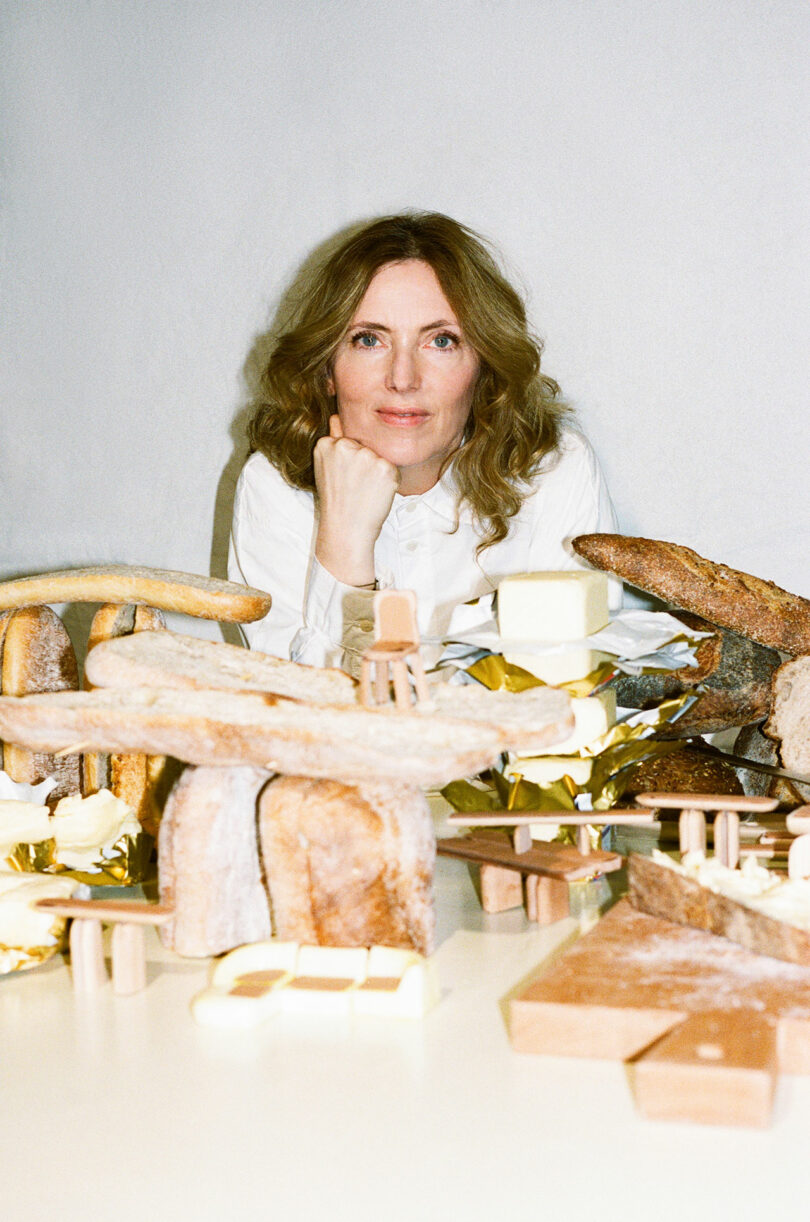 A woman with wavy hair and a white shirt sits at a table that has bread, cheese and other foods arranged in sculptural forms reminiscent of Faye Toogood's aesthetics.