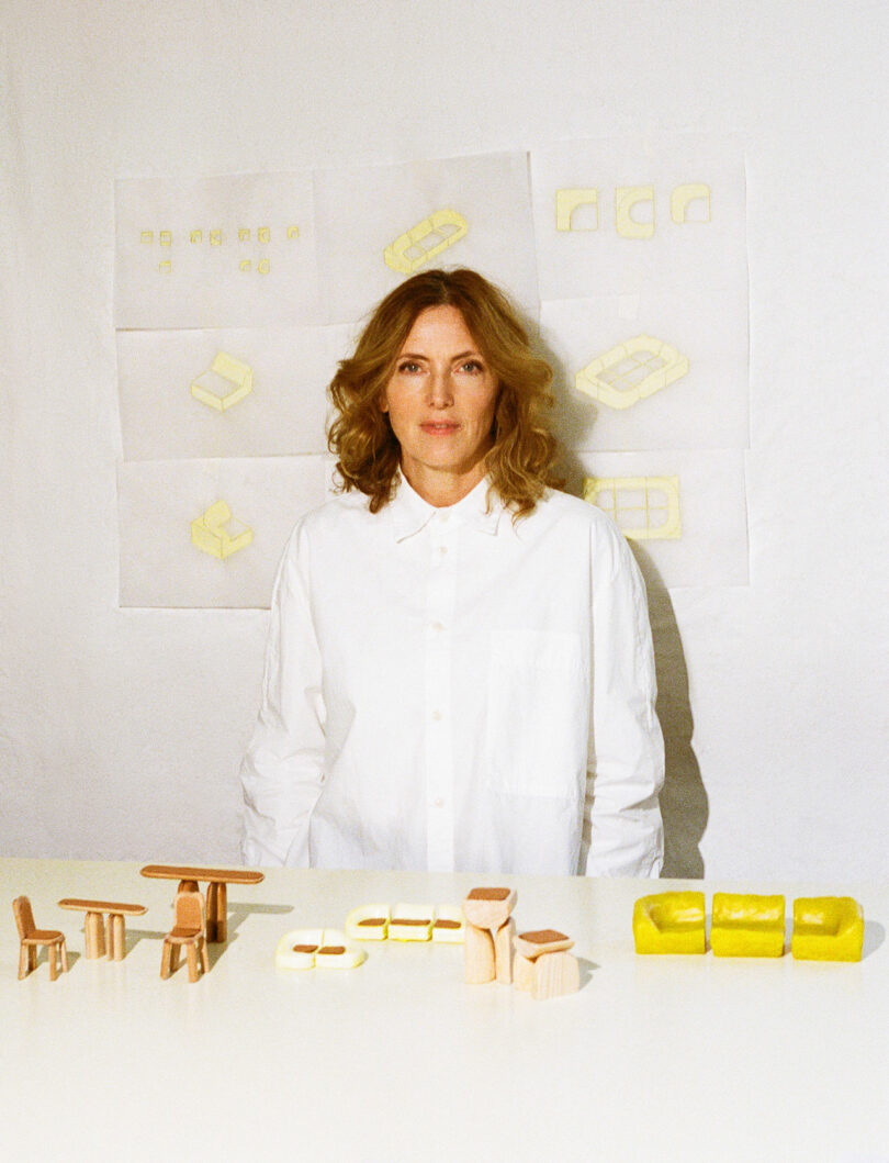 A person in a white shirt is behind a table that has small models of wood and yellow, with sketches of Faye Toogood furniture on the wall behind them.