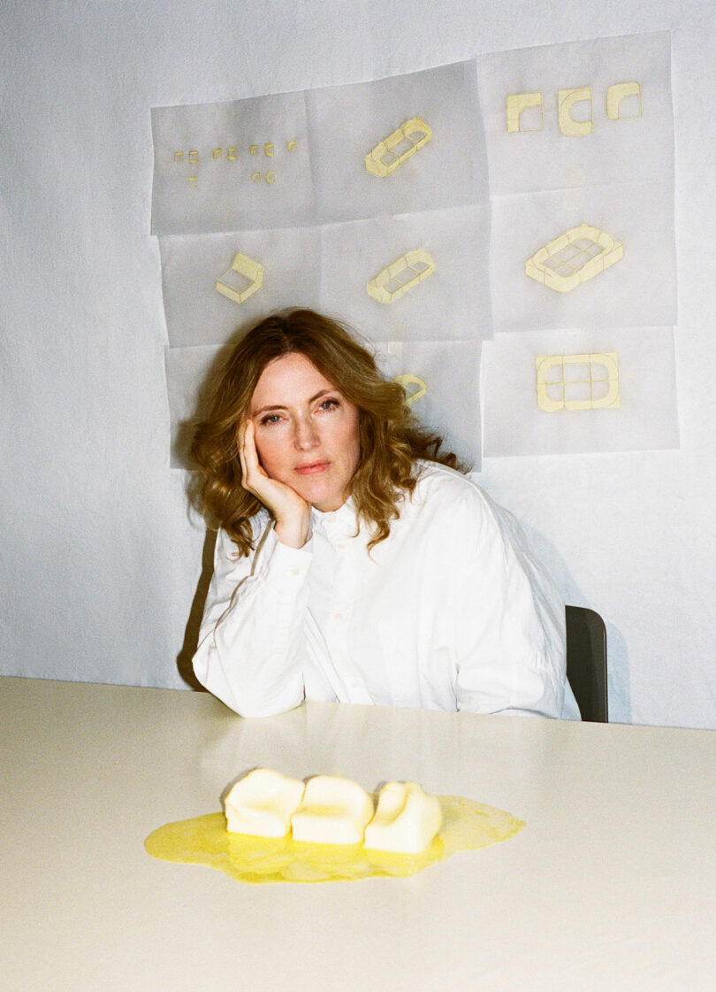 A woman in a white shirt sits at a melting butter in front of her; Behind her, butter sketches - reminiscent of Faye Toogood's artistic style - are lined on the wall.