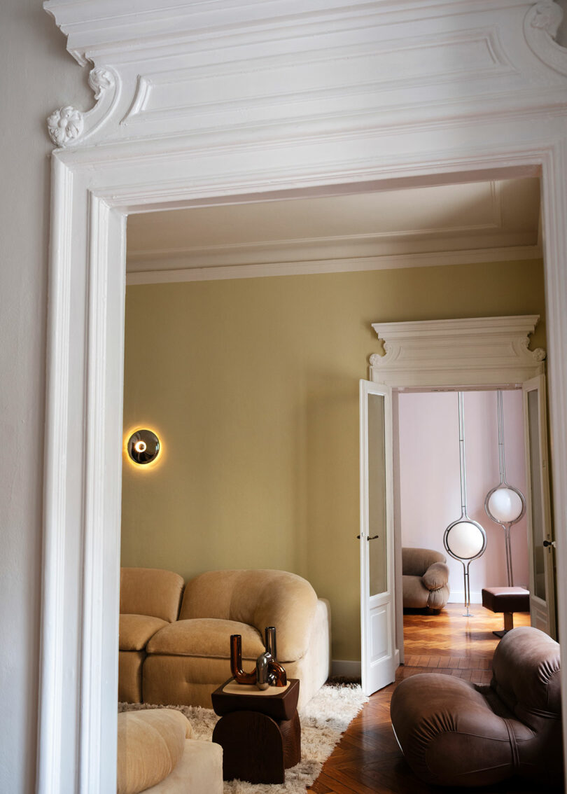 A view through the white doors ornamented in a living room with beige curved sofas, inspired by Faye Toogood, a round table, soft carpet and modern circular wall lights and floor.