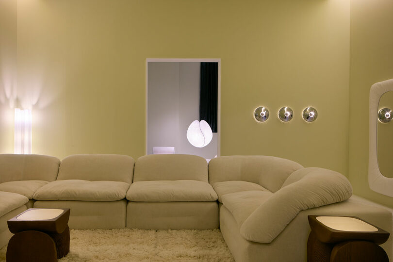 A modern living room, with a large beige sectional sofa, shaggy carpet, two wooden lateral tables inspired by Faye Toogood, soft lighting and round wall application on a pale green wall.